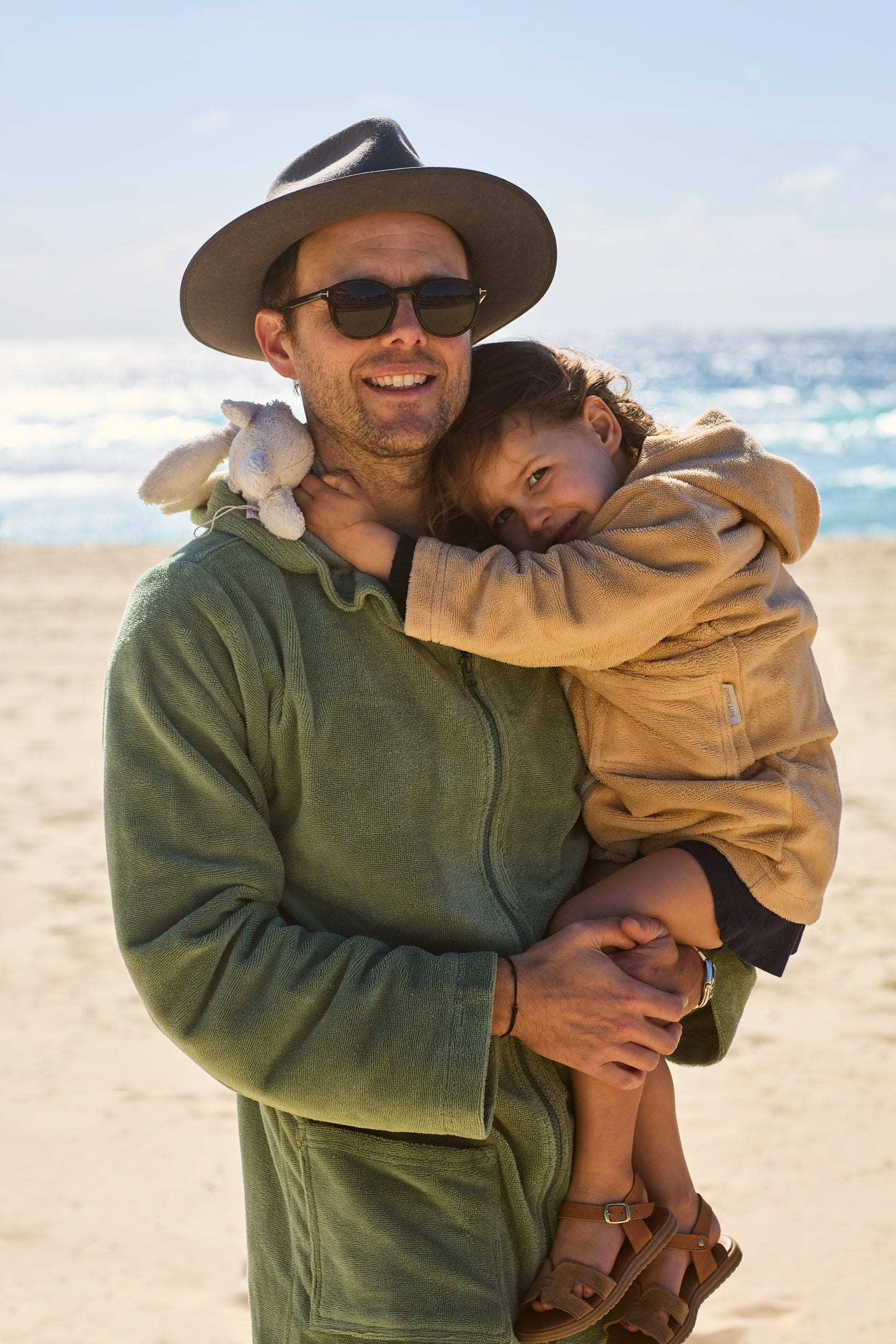 Toddler wearing a Sandcastle hooded towel from Capimiri brand in her dad's arms by the beach.