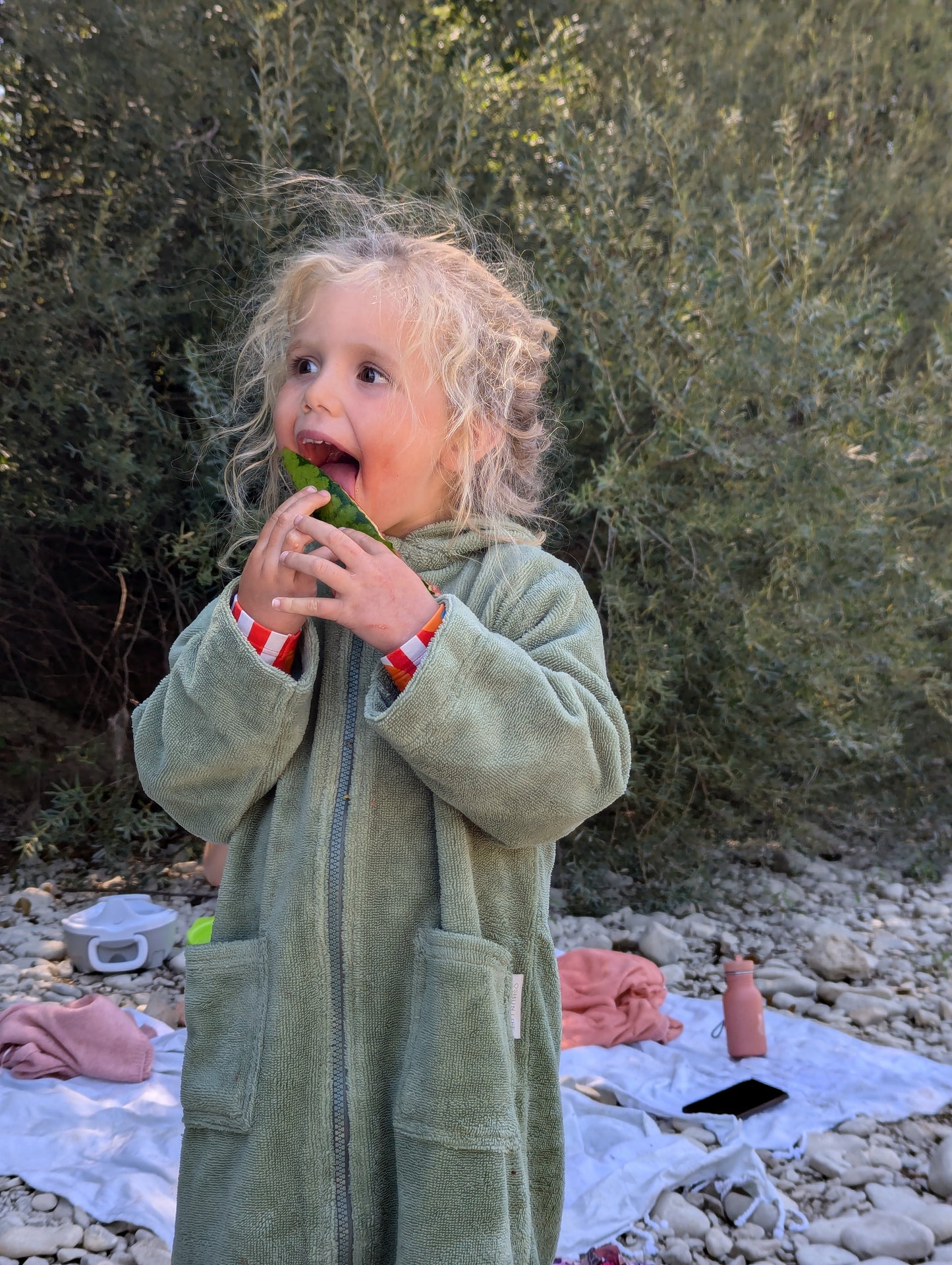 Child in a green towel eating watermelon outdoors with bushes in the background