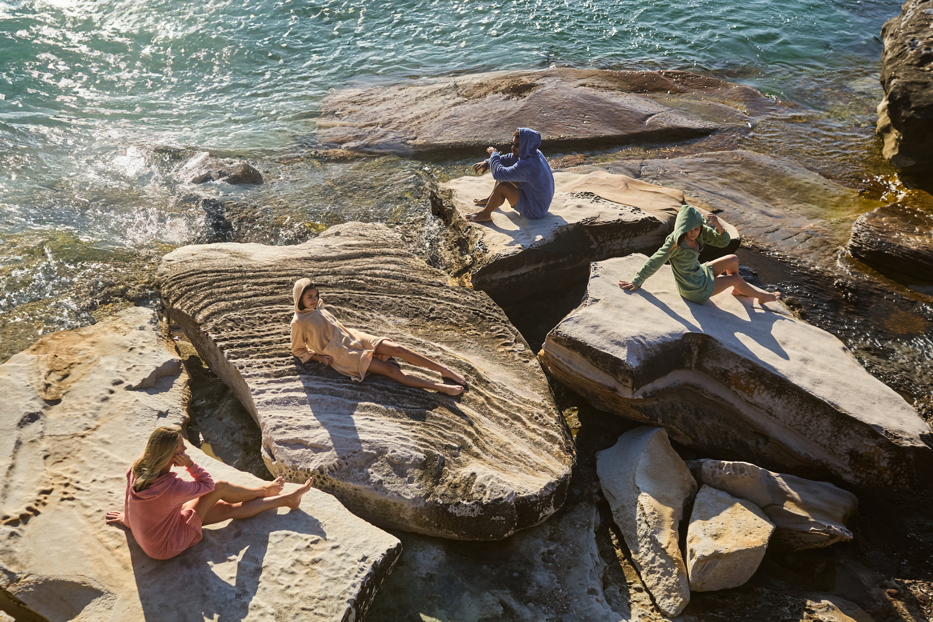 Models wearing CAPIMIRI hooded towels on the rocks by the ocean.