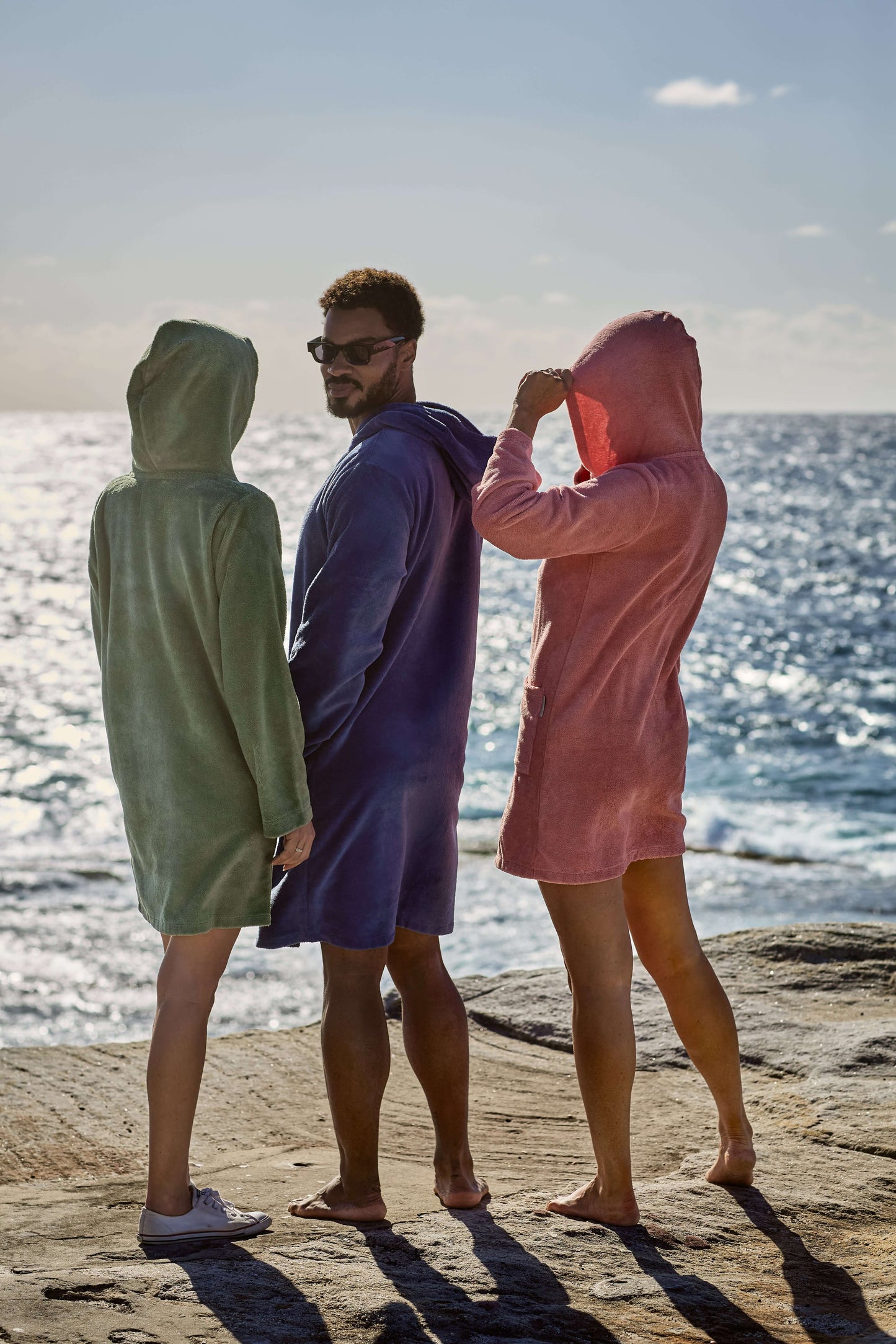 A man and two women chilling by the ocean, with their CAPIMIRI garment on. Guava Women's Hooded Towel