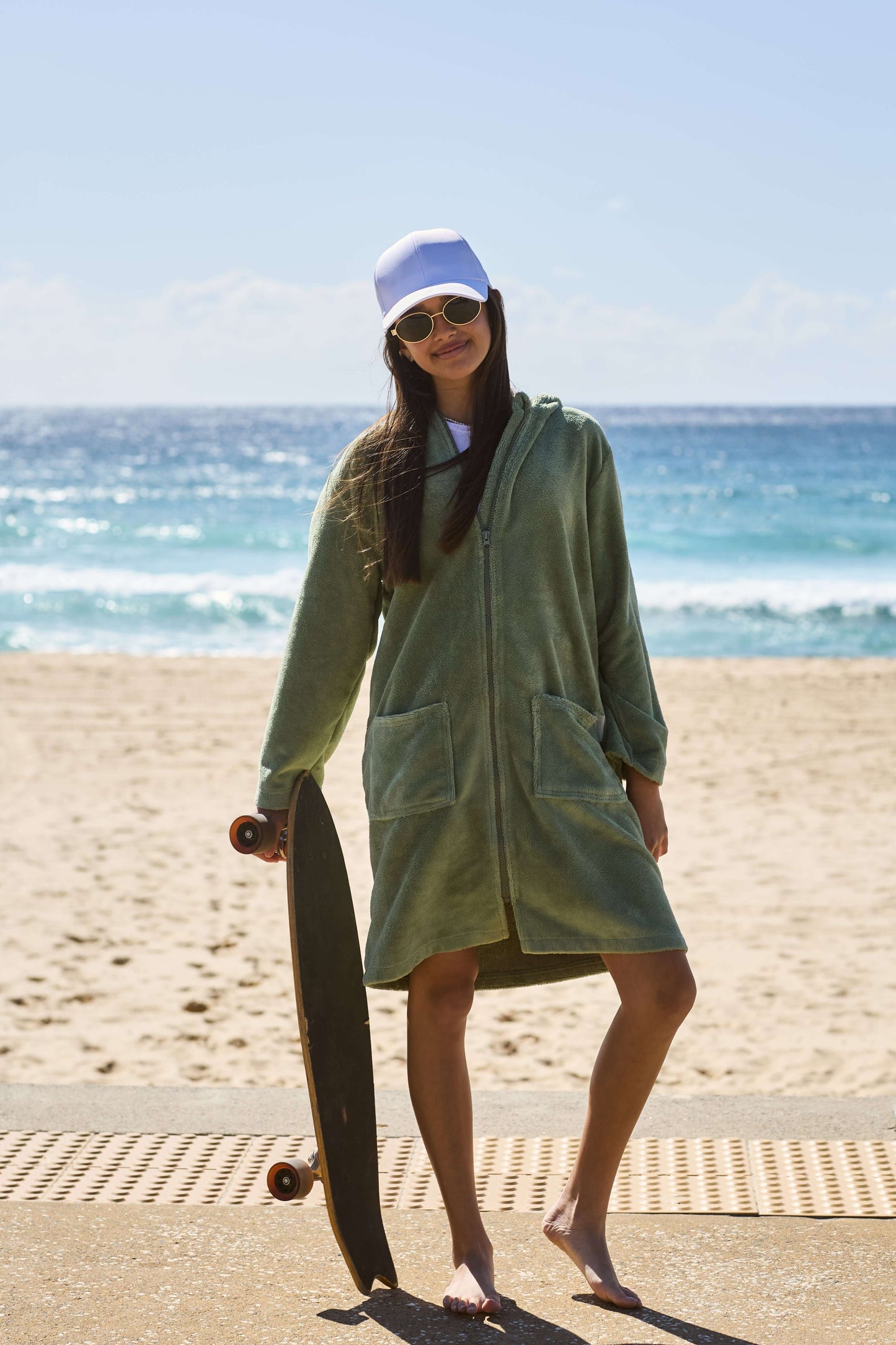Young girl wearing a green hooded towel with her skateboard in one hand, barefoot by the ocean. Cactus Pre Teens & Teens