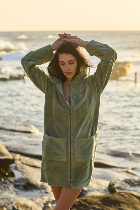 Model wearing a CAPIMIRI Cactus beach hooded towel at sunrise with rocks and ocean pool around. This soft and elegant garment is a must have in your wardrobe