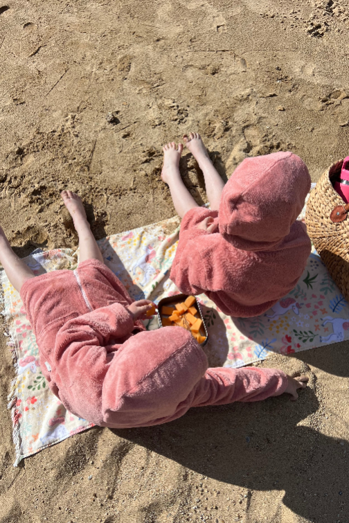 kids in pink towel sitting on a beach with a colorful blanket and snacks