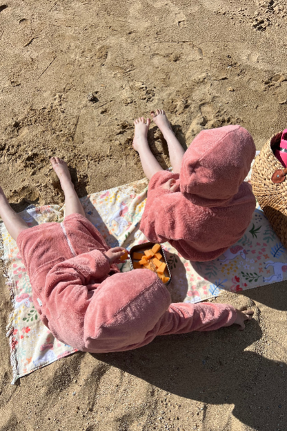 kids in pink towel sitting on a beach with a colorful blanket and snacks