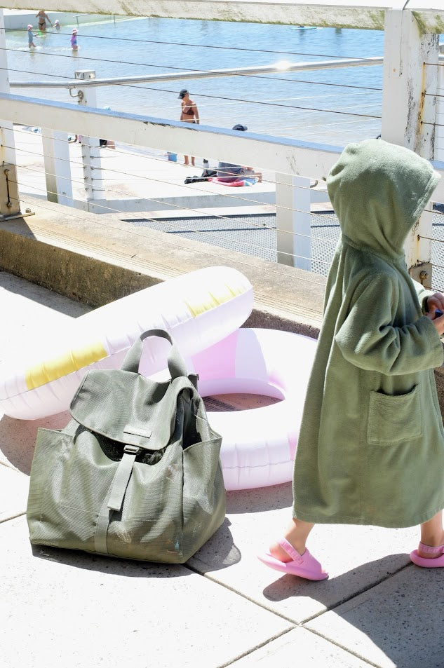 Person in a green robe standing next to a green bag on a tiled floor with a beach and pool area in the background.